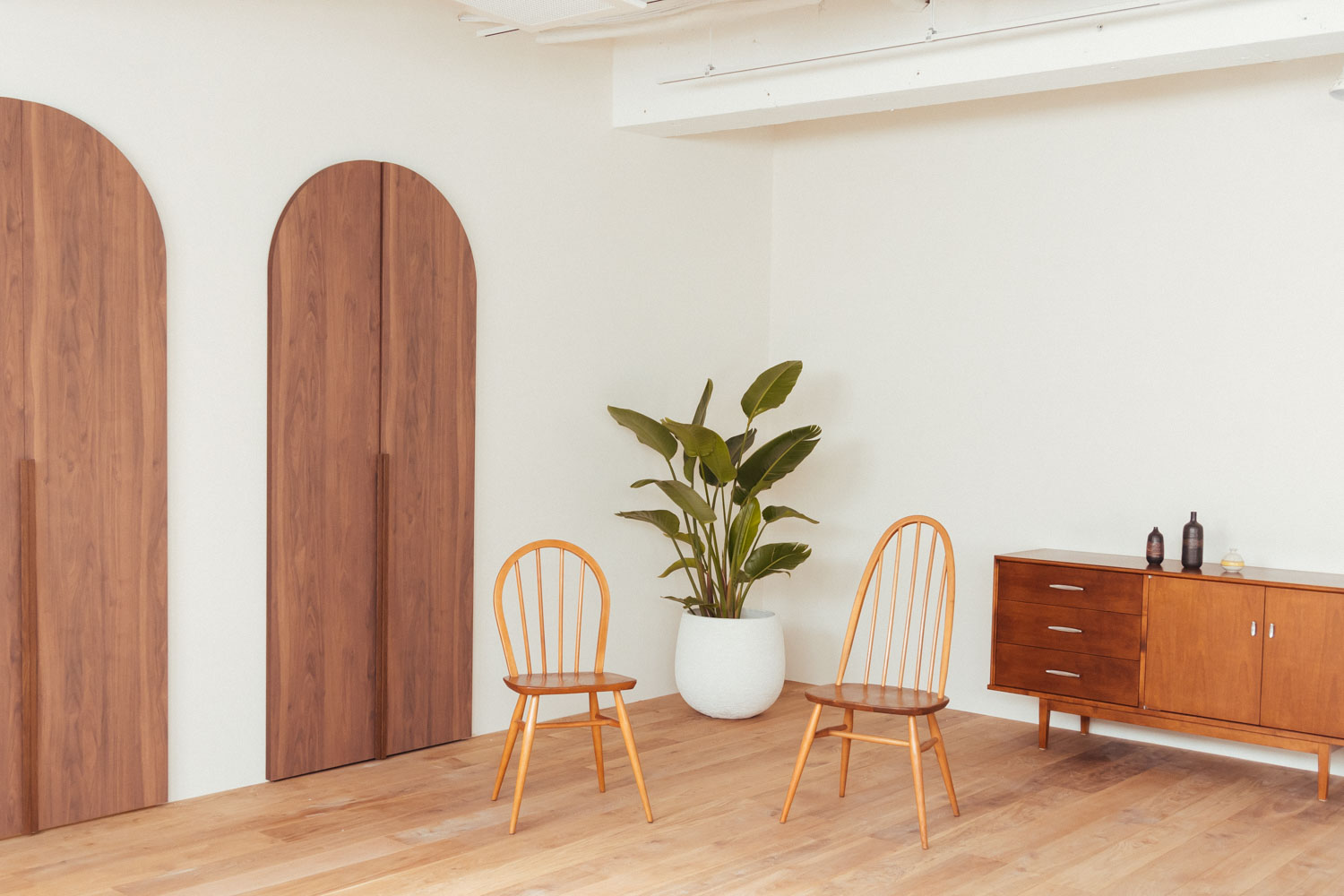 円形の木製ドアがあるフローリングの部屋に、アーコールチェア2脚と観葉植物、チェストが配置された写真